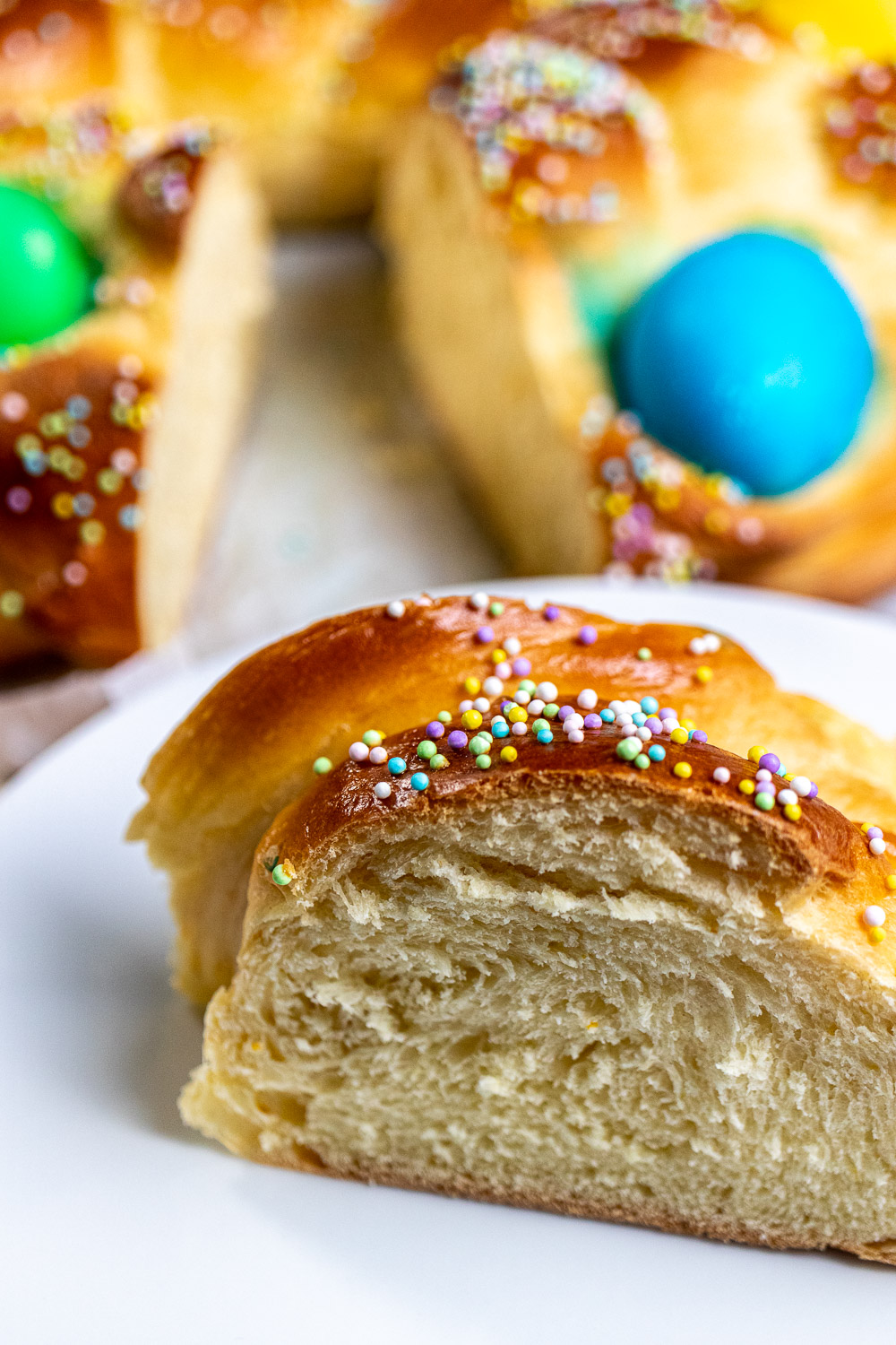 close up view of a slice of Italian Easter bread with dyed eggs and sprinkles with a full loaf behind