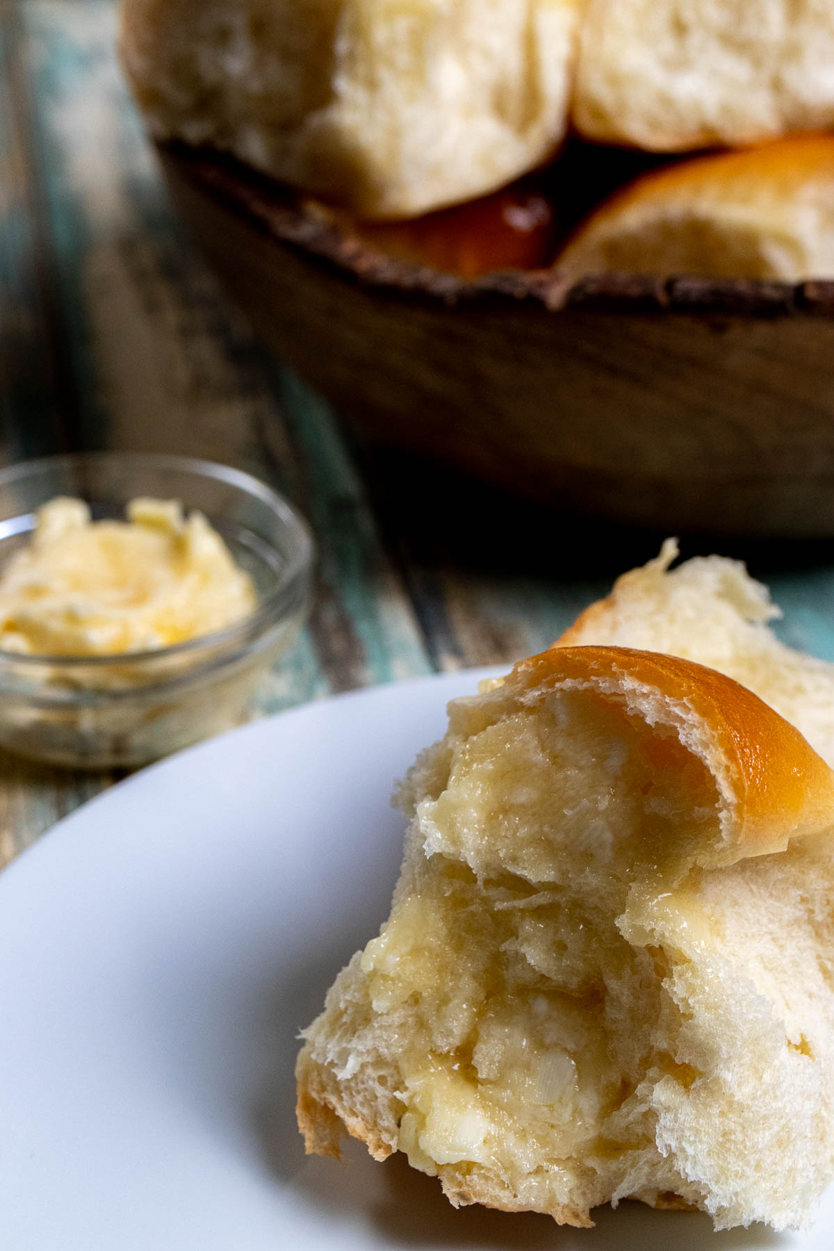 Honey butter dinner rolls in a wooden bowl with a side of honey butter in a small dish. A split open roll in a white plate slathered with honey butter in the foreground. 