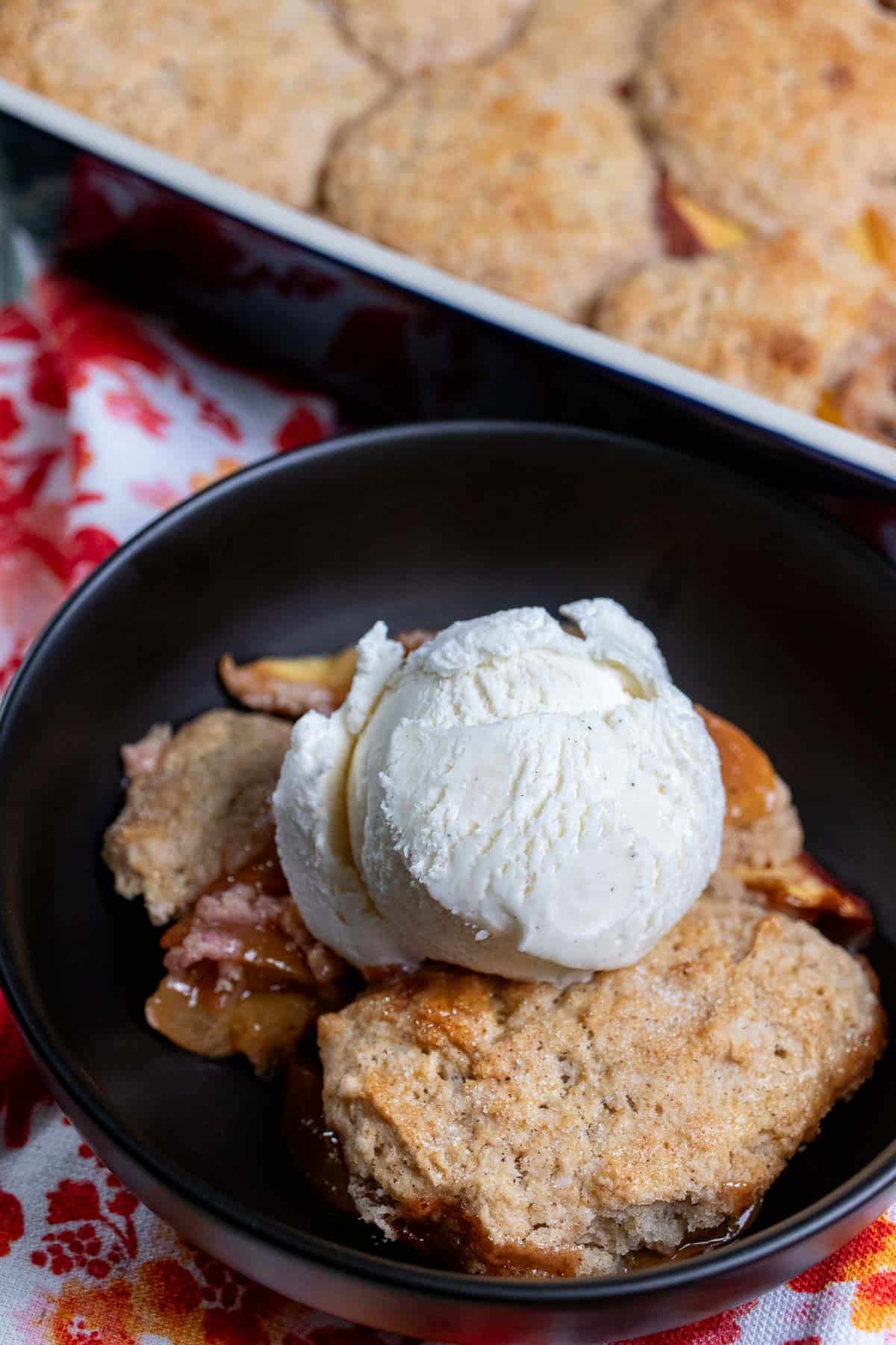 Biscuit topped peach cobbler in a baking dish, with a black bowl of peach cobbler topped with ice cream in front.