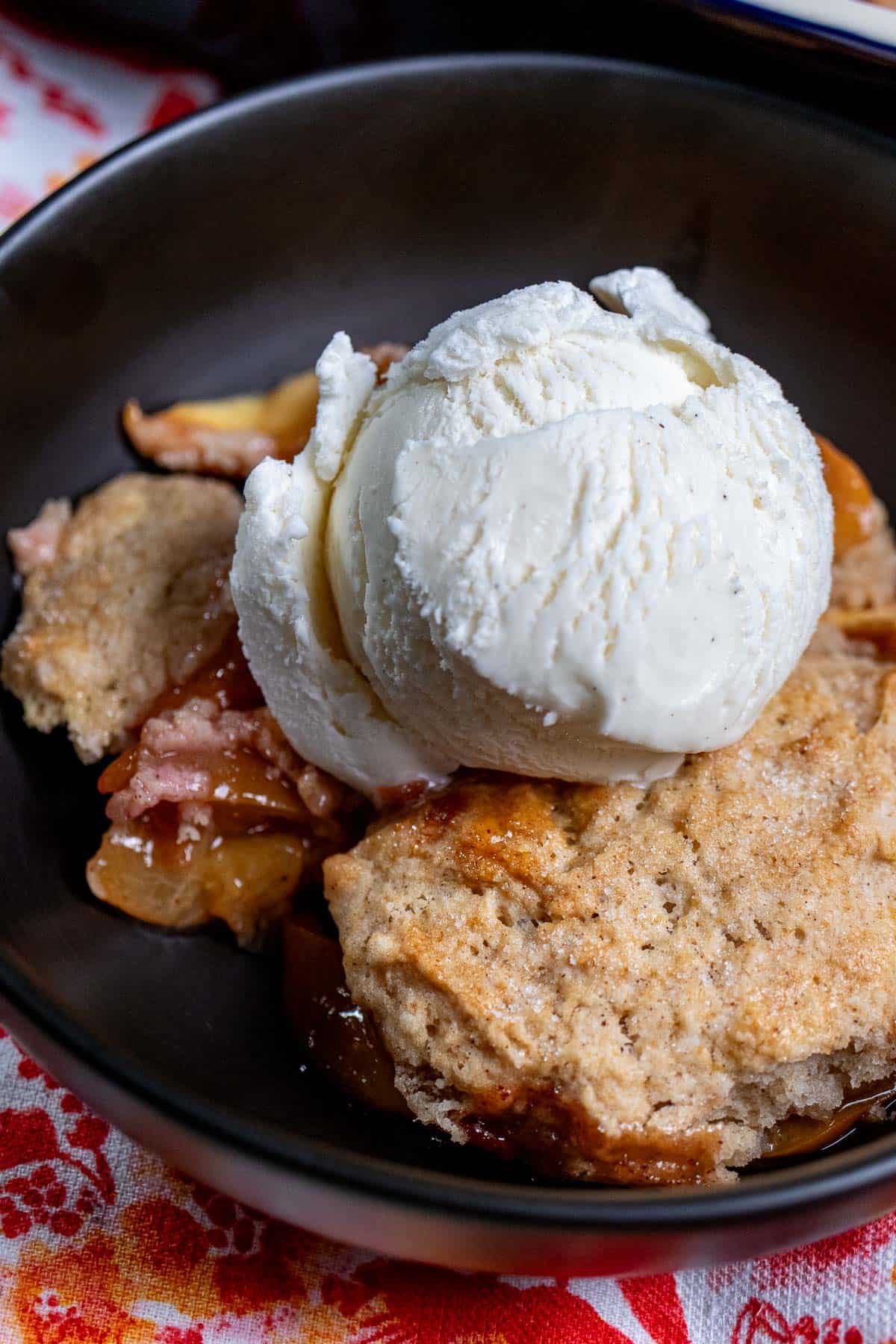 Close up view of fresh peach cobbler in a black bowl, topped with vanilla ice cream.