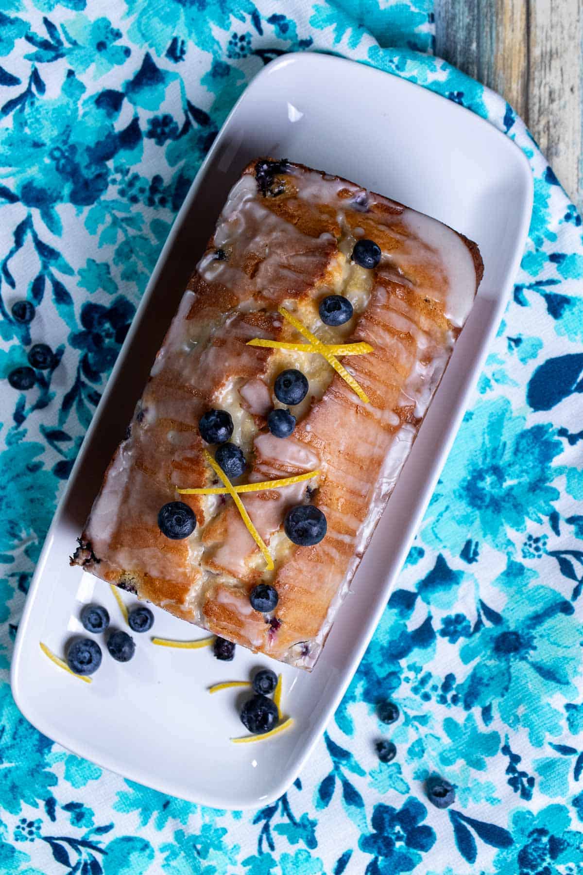Overhead view of a glazed lemon blueberry pound cake on a white plate with blueberries and lemon zest around it.