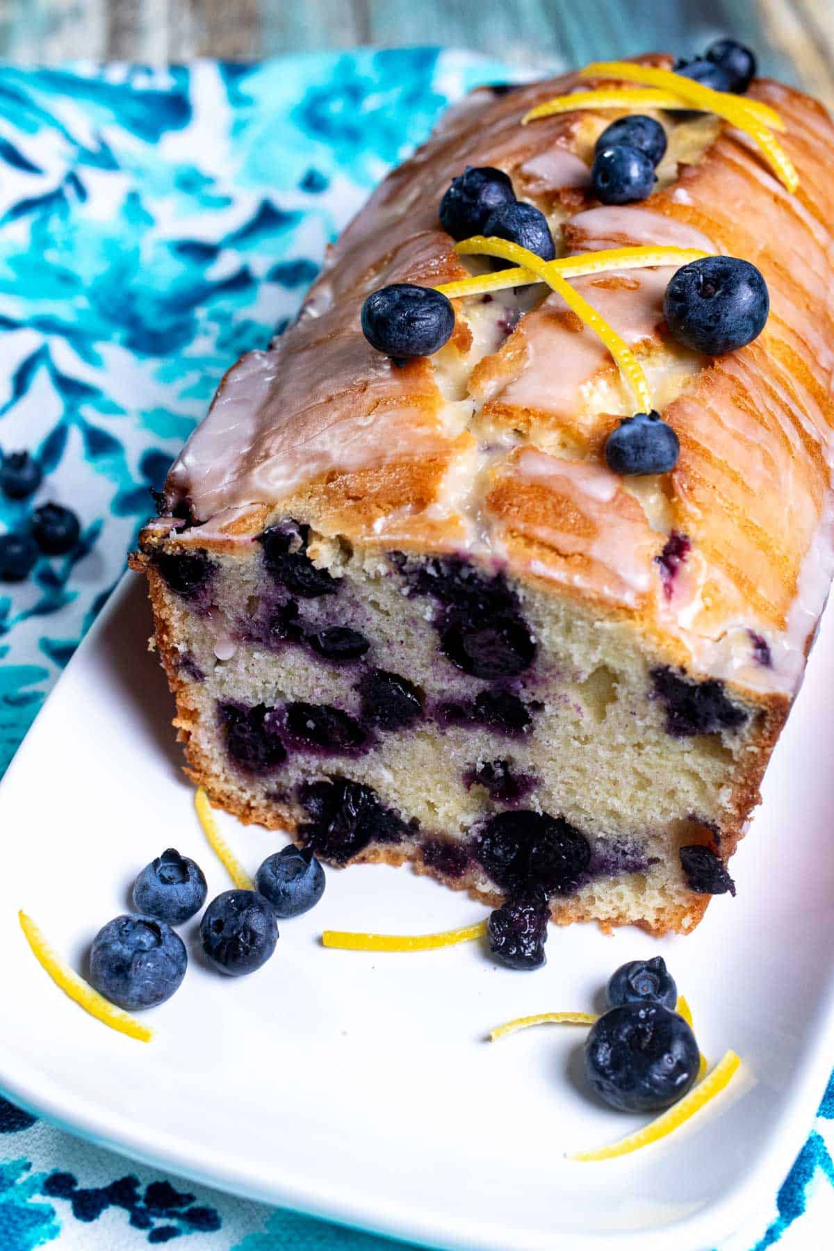 Angled view of a sliced, glazed, lemon blueberry pound cake on a white plate with blueberries and lemon zest around it.