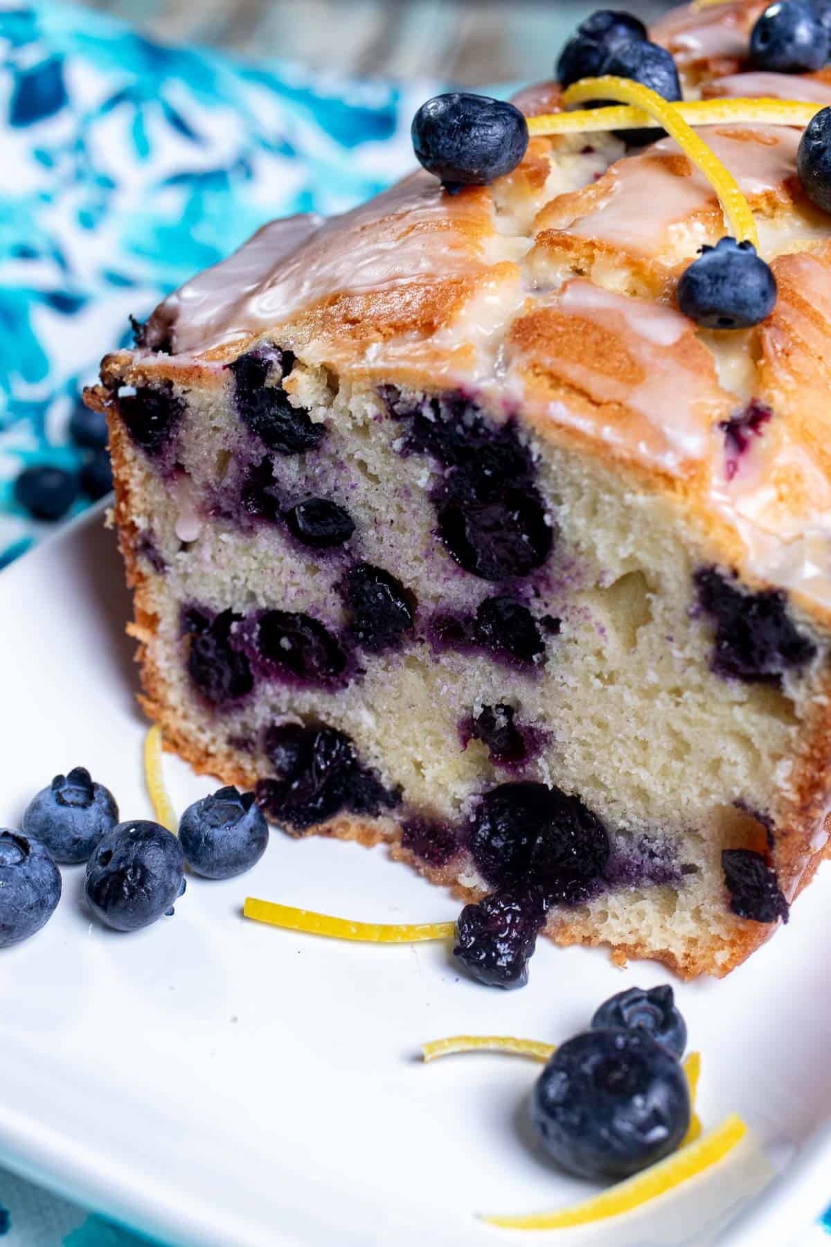 Close up view of a sliced, glazed, lemon blueberry pound cake on a white plate with blueberries and lemon zest around it.