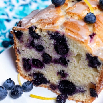 Close up view of a sliced, glazed, lemon blueberry pound cake on a white plate with blueberries and lemon zest around it.