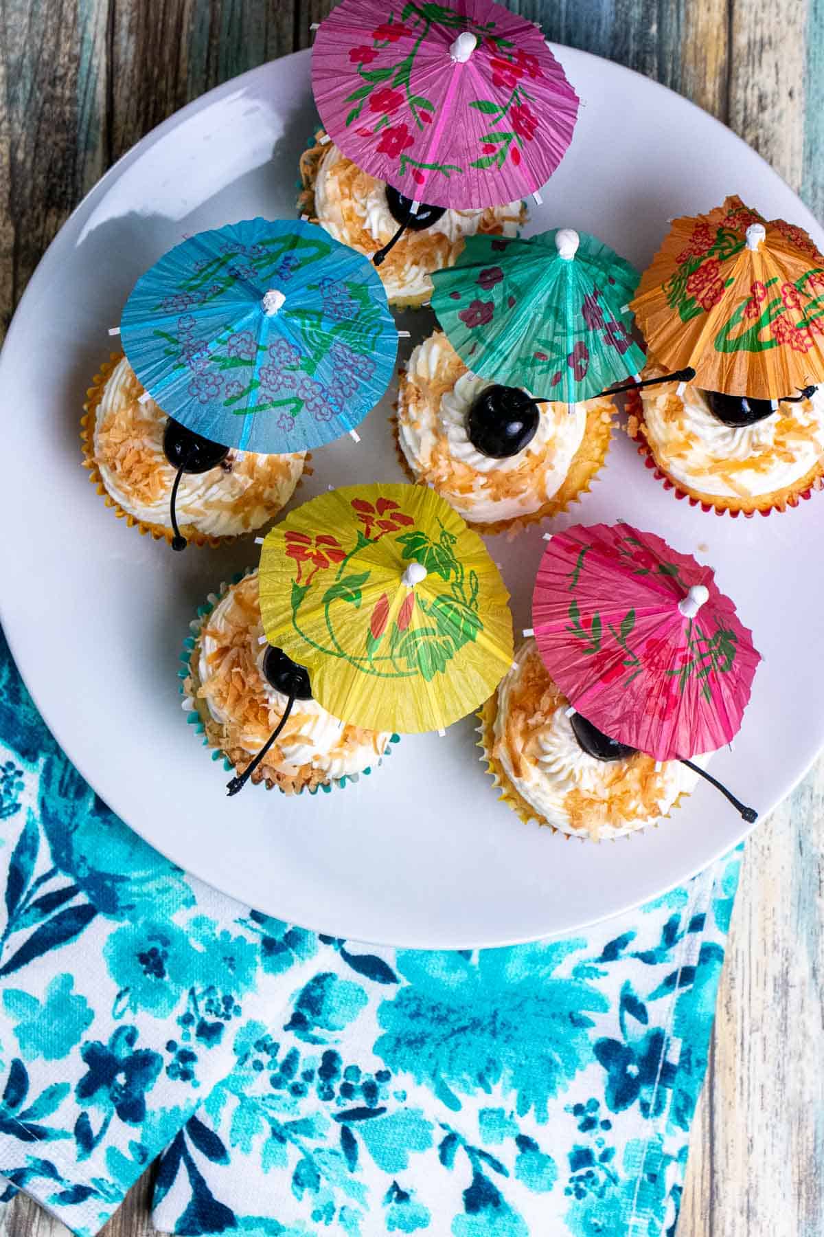 Overhead view of pina colada cupcakes on a white plate over a blue towel, decorated with toasted coconut, a cherry, and a cocktail umbrella.