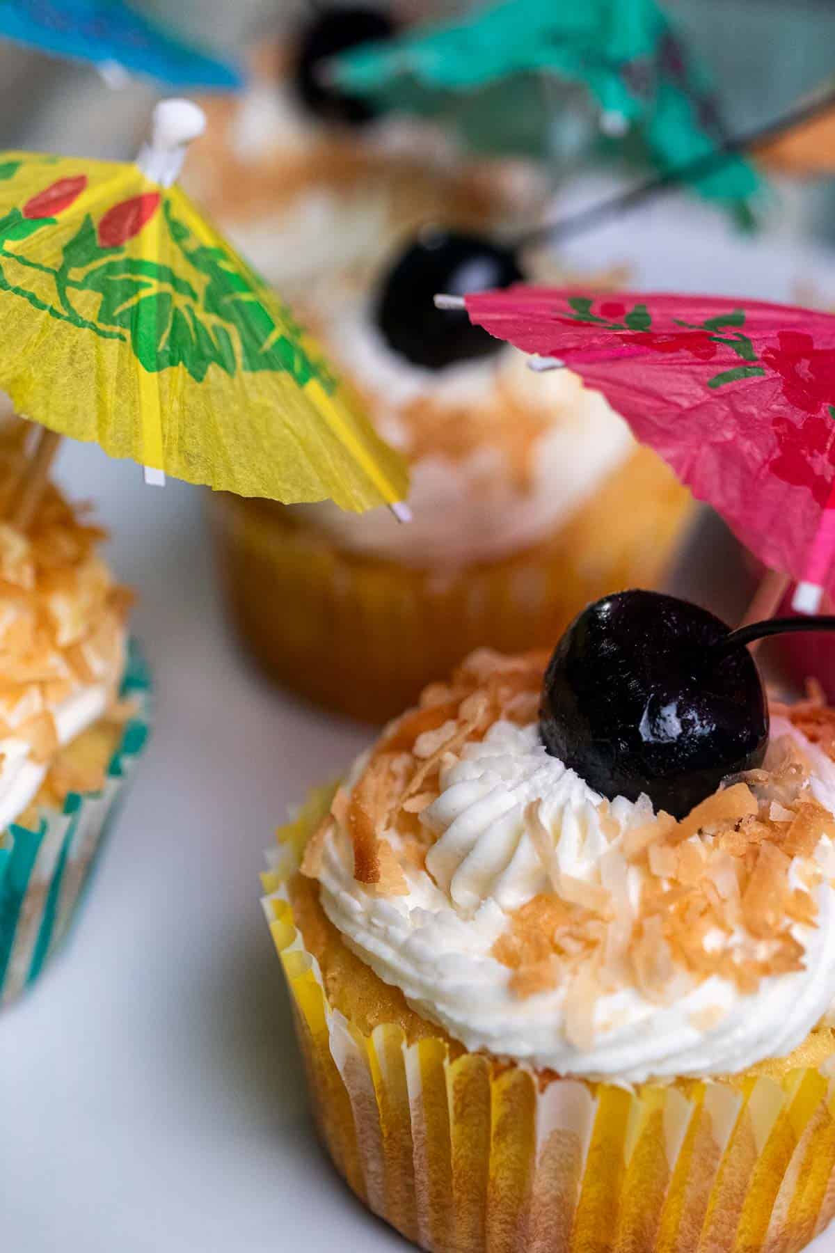 Close up of pina colada cupcakes decorated with toasted coconut, a cherry, and a cocktail umbrella.