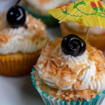 Close up of pina colada cupcakes decorated with toasted coconut, a cherry, and a cocktail umbrella.