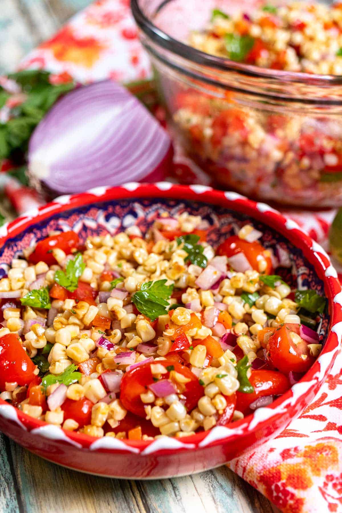 Closer view of grilled corn salad in a red and white bowl with half a red onion and parsley behind it.