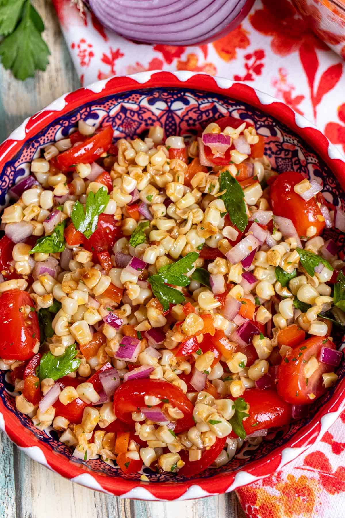 Close up view of grilled corn salad in a red and white bowl with half a red onion and parsley behind it.