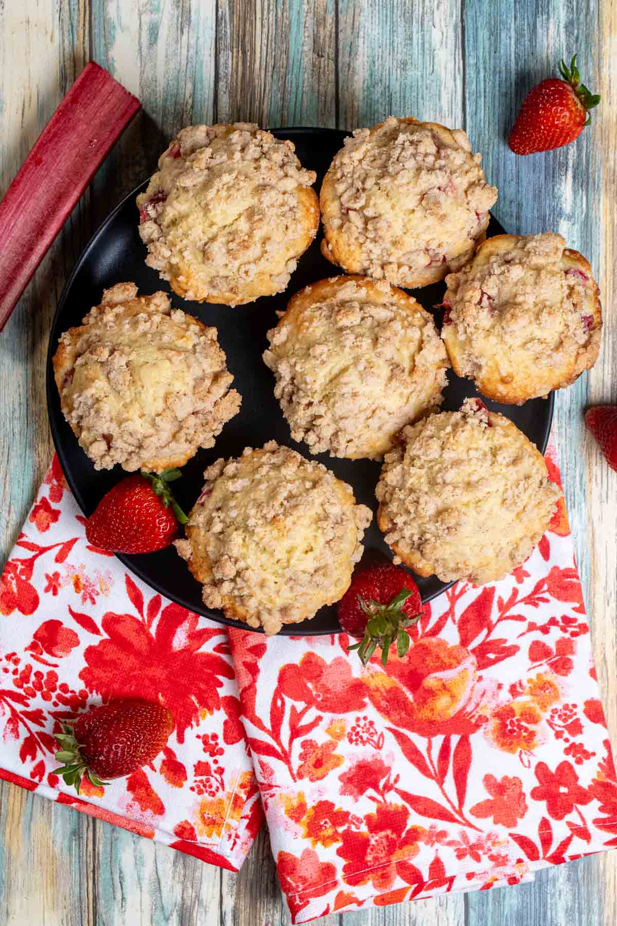 Overhead view of strawberry rhubarb streusel muffins on a black plate with strawberries and a piece of rhubarb around them. 