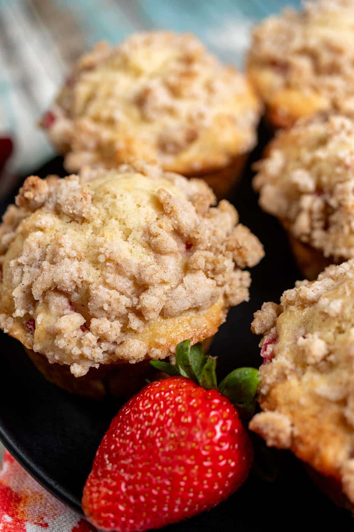 Close up view of strawberry rhubarb streusel muffins on a black plate with a large, red strawberry next to them.