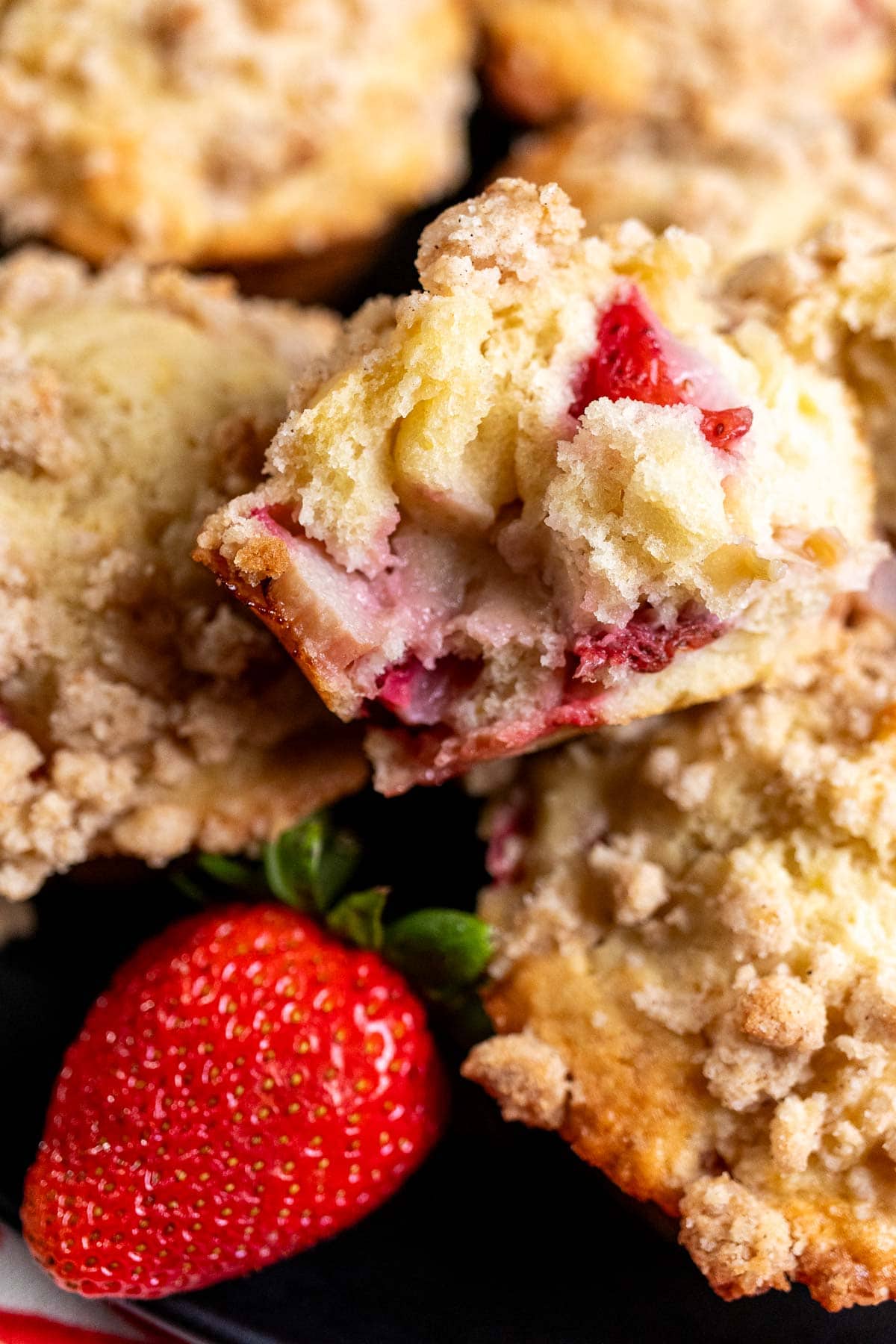 Strawberry rhubarb streusel muffins on a black plate with a large, red strawberry next to them and a muffin cut open on top of the others.