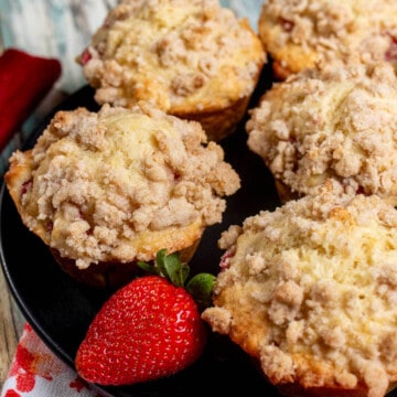 Strawberry rhubarb streusel muffins on a black plate with a large, red strawberry next to them.