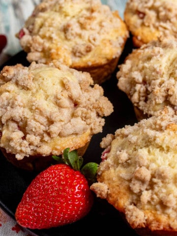 Strawberry rhubarb streusel muffins on a black plate with a large, red strawberry next to them.