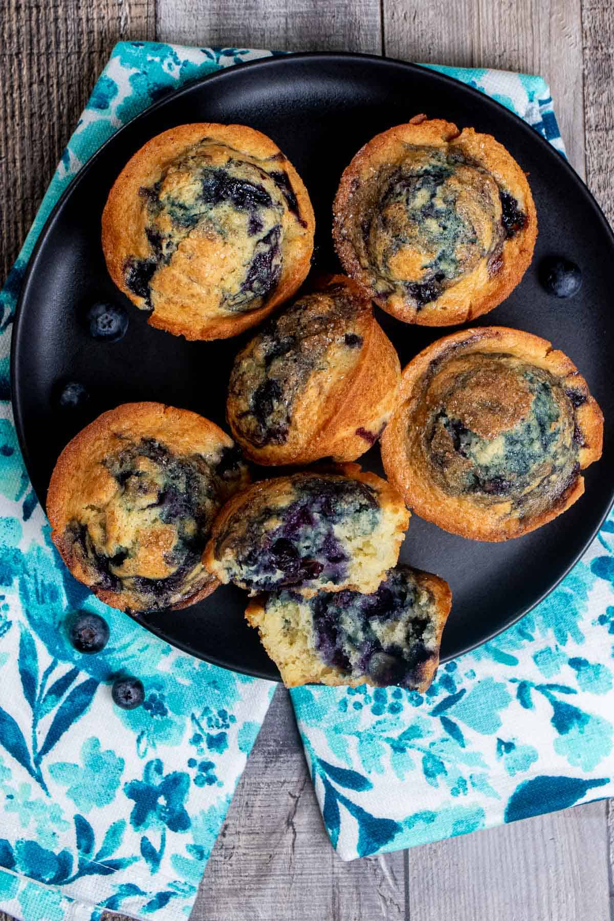 Overhead view of a black plate on a blue and white cloth with a pile of blueberry swirl muffins on top.