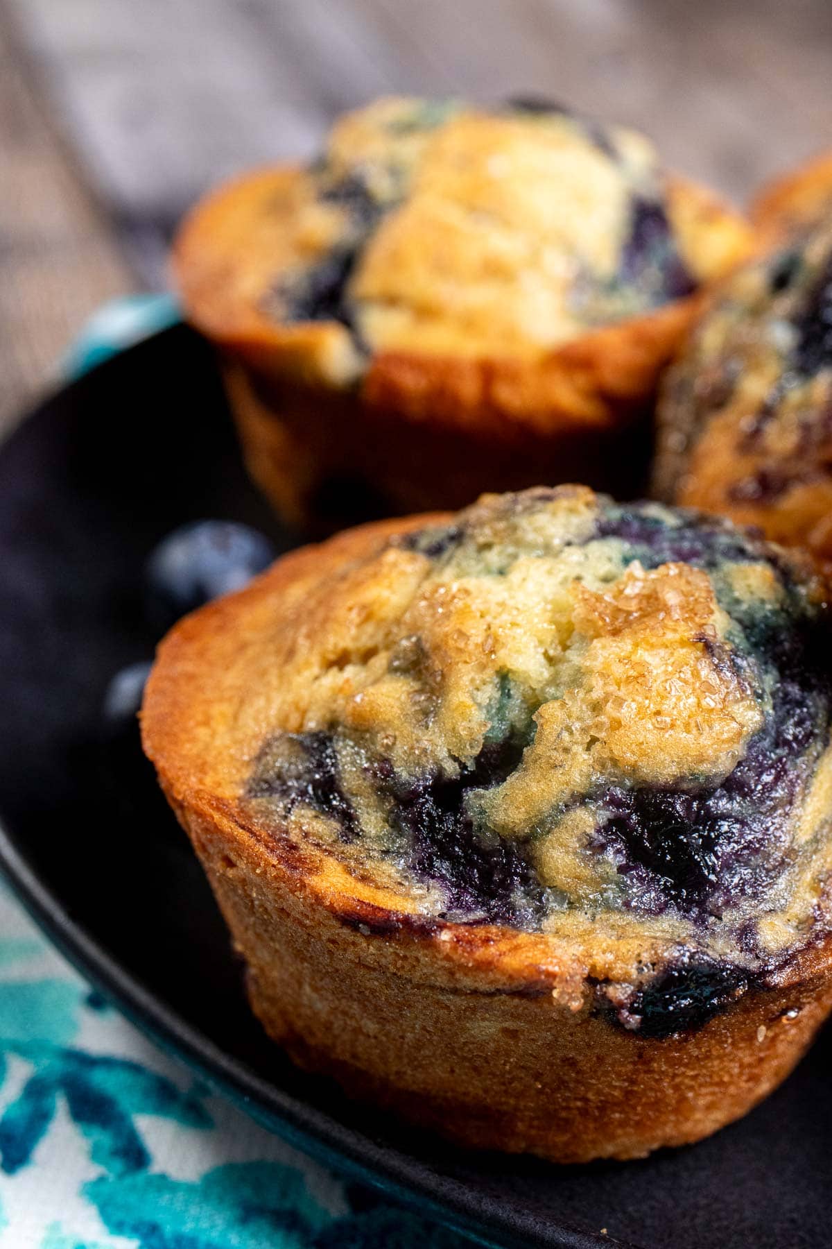 Side view of a black plate on a blue and white cloth with a pile of blueberry swirl muffins on top.