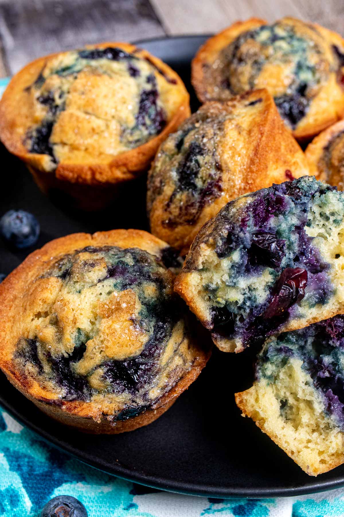 Bakery style blueberry swirl muffins on a black plate on top of a blue and white cloth. One muffin is cut open to show the swirl of blueberries inside. 