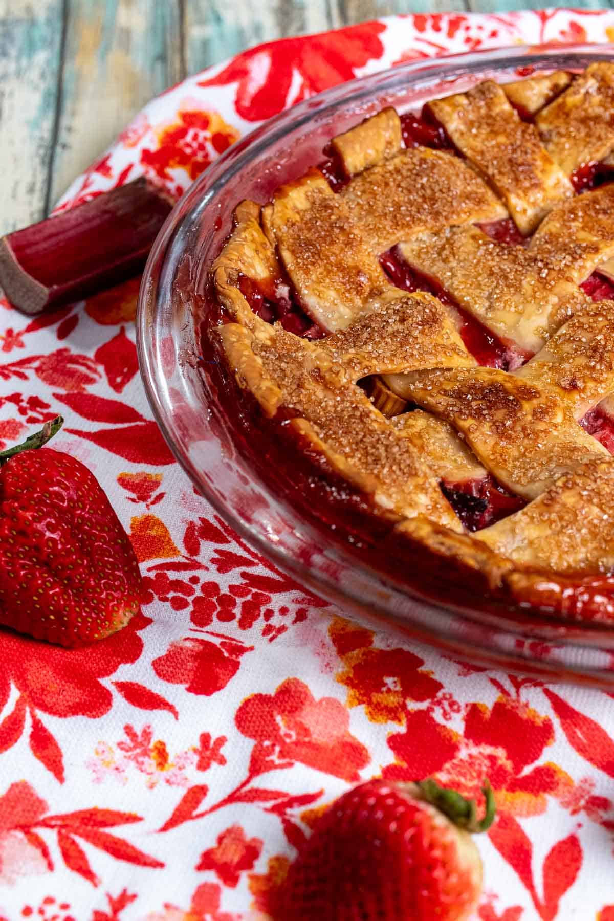 Strawberry rhubarb pie in a glass dish on top of a red and white floral cloth, with strawberries around it. 