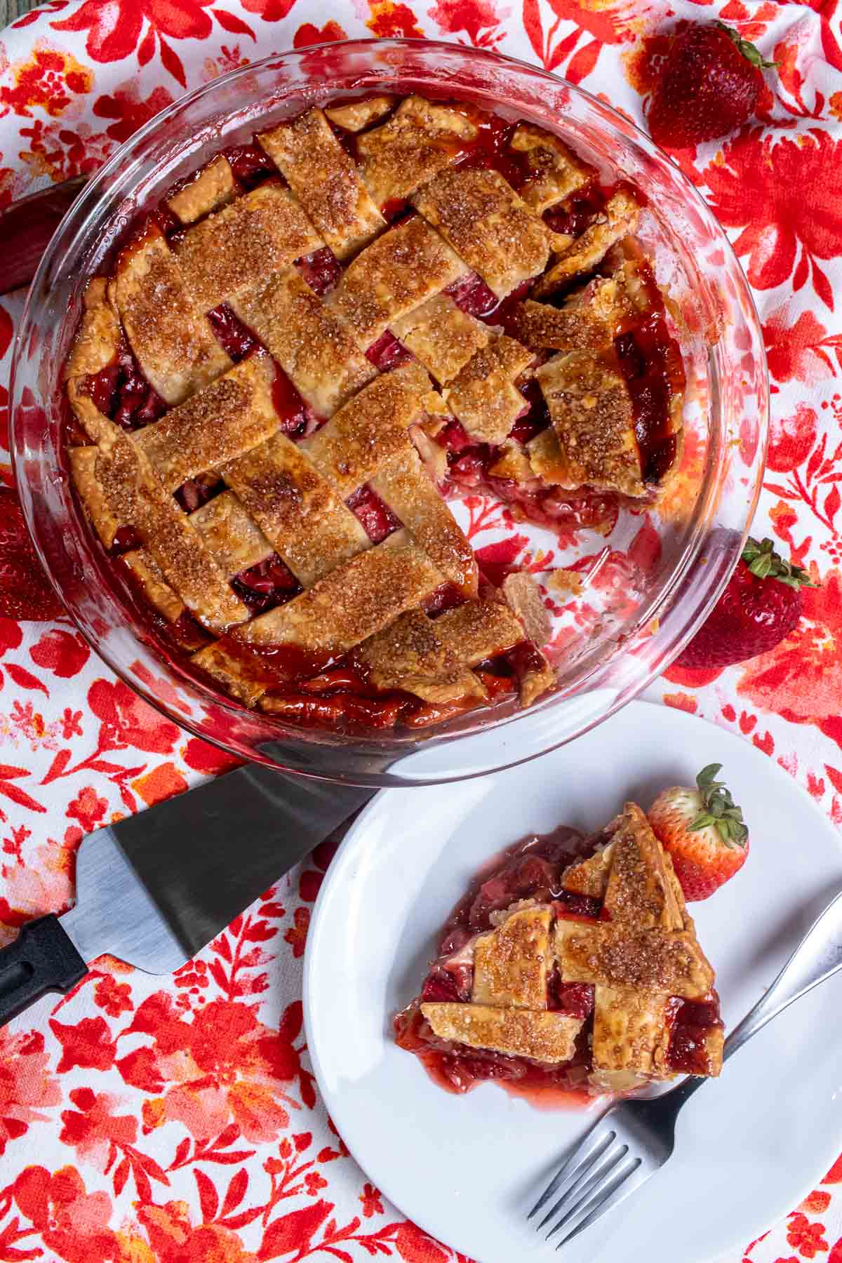 Overhead view of strawberry rhubarb lattice pie on a red and white floral cloth. One slice removed and on a white plate in front of the pie. 