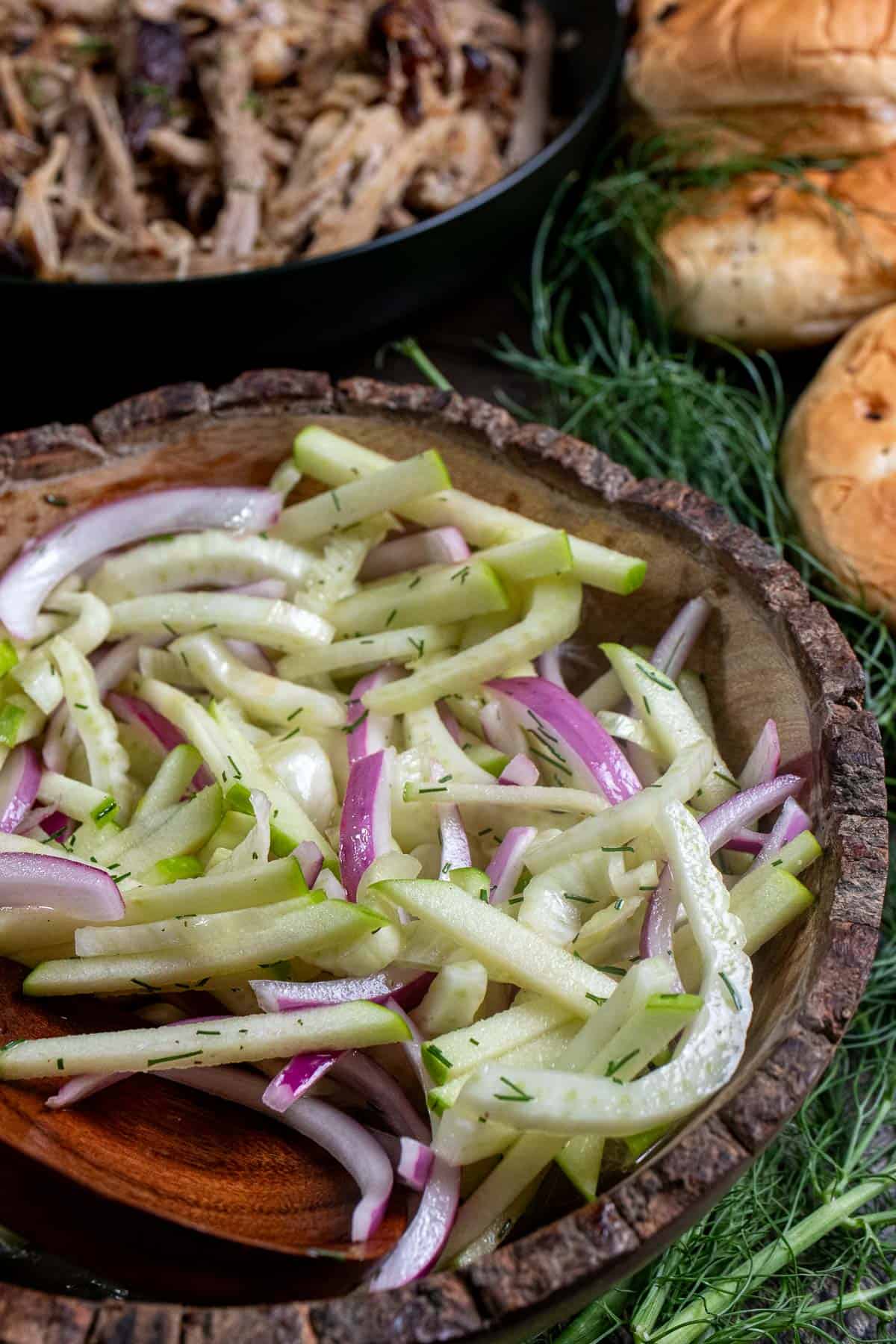 Fennel and apple slaw in a wooden bowl with a plate of cider pulled pork behind it.