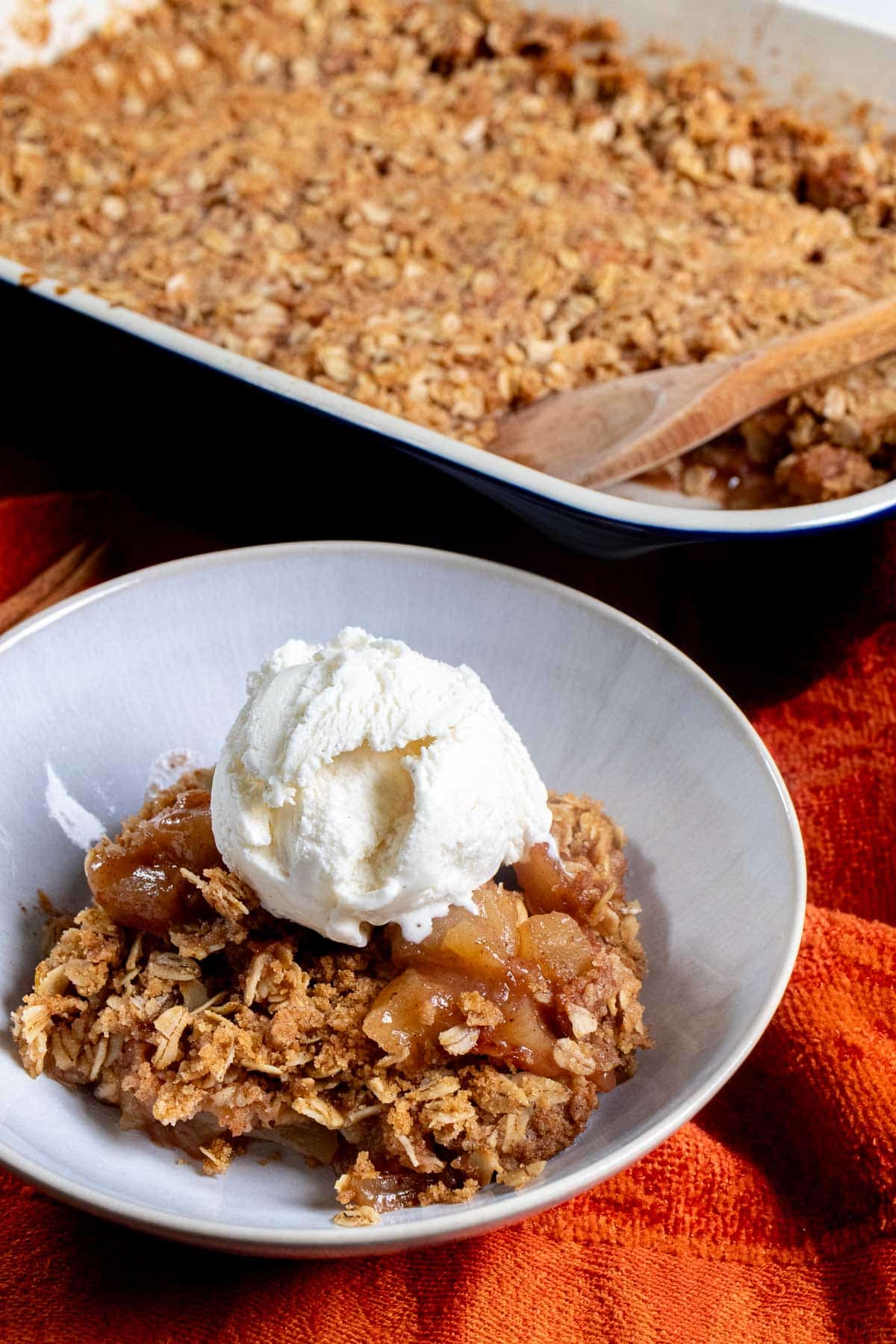 Angled view of an apple crisp in a 13x9 dish with a portion in a white bowl next to it, topped with ice cream.