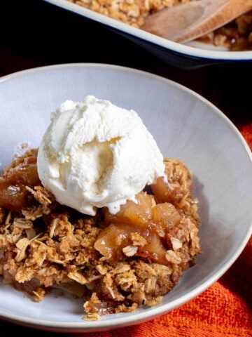 Angled view of a portion of apple crisp in a white bowl, topped with vanilla ice cream.