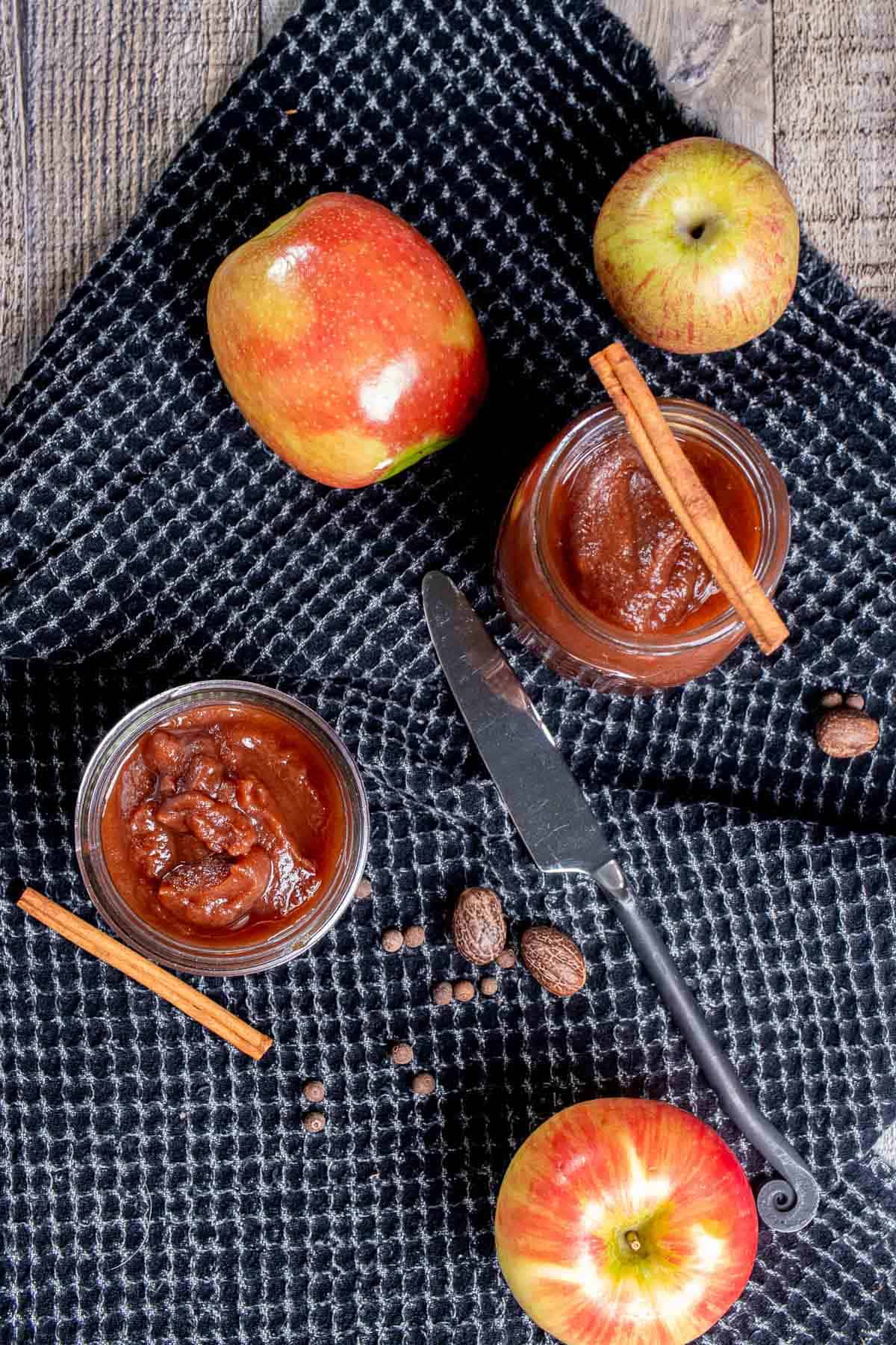 Overhead view of jars of apple butter on a black cloth surrounded by apples, cinnamon sticks, and whole nutmegs.