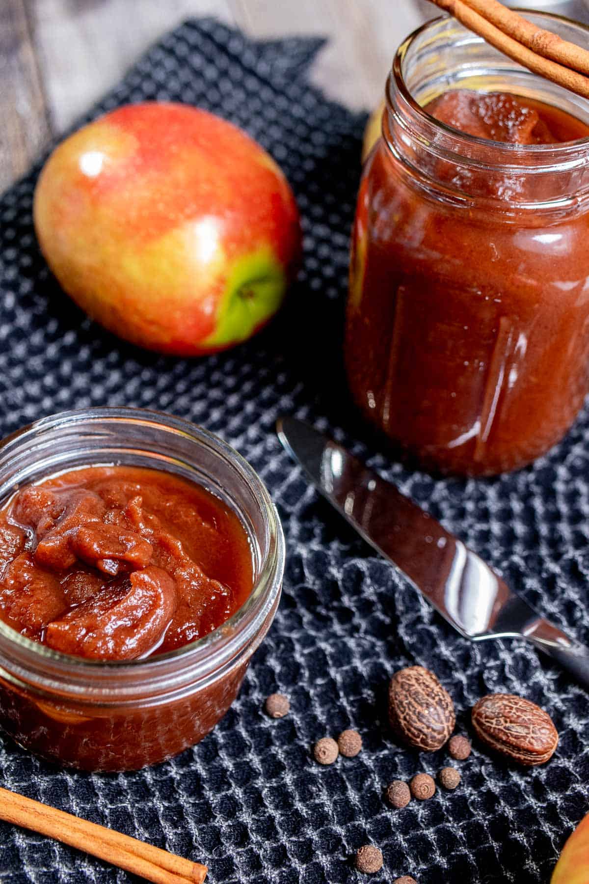 Angled view of jars of apple butter on a black cloth surrounded by apples, cinnamon sticks, and whole nutmegs.