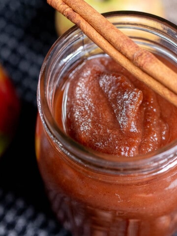 Close up view of apple butter in a pint jar with a cinnamon stick laid across the top of the open jar.