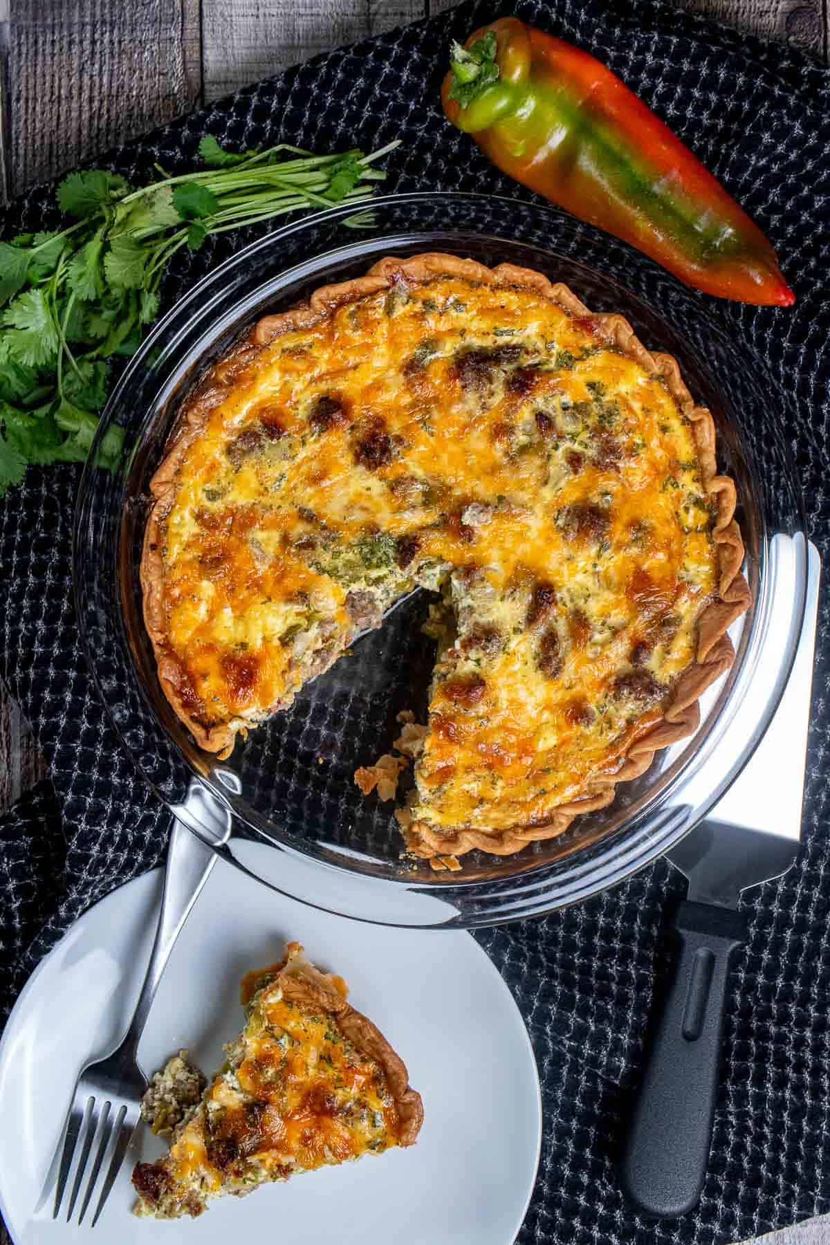 Overhead view of Hatch chile sausage quiche in a glass pie dish with a slice on a white plate in front of it.
