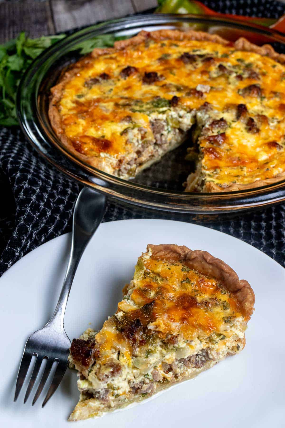 Angled view of Hatch chile sausage quiche in a glass pie dish with a slice on a white plate in front of it.