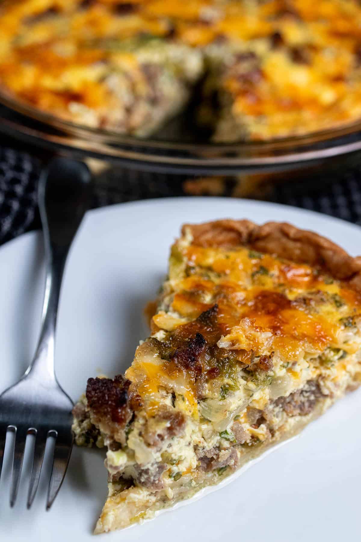Angled view of green chile and sausage quiche in a glass pie dish with a slice on a white plate in front of it.