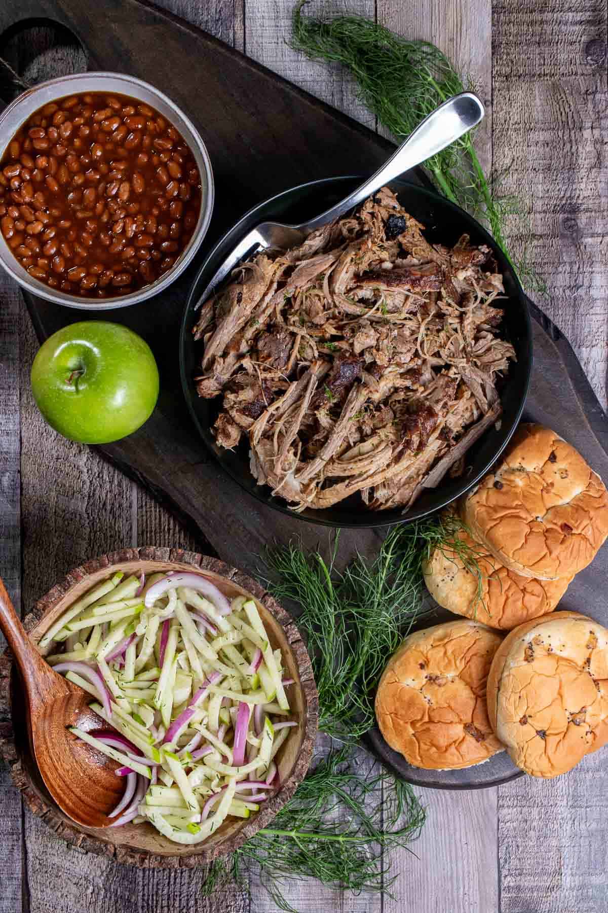 Overhead view of cider braised pork shoulder, served with onion buns, fennel apple slaw, and baked beans.