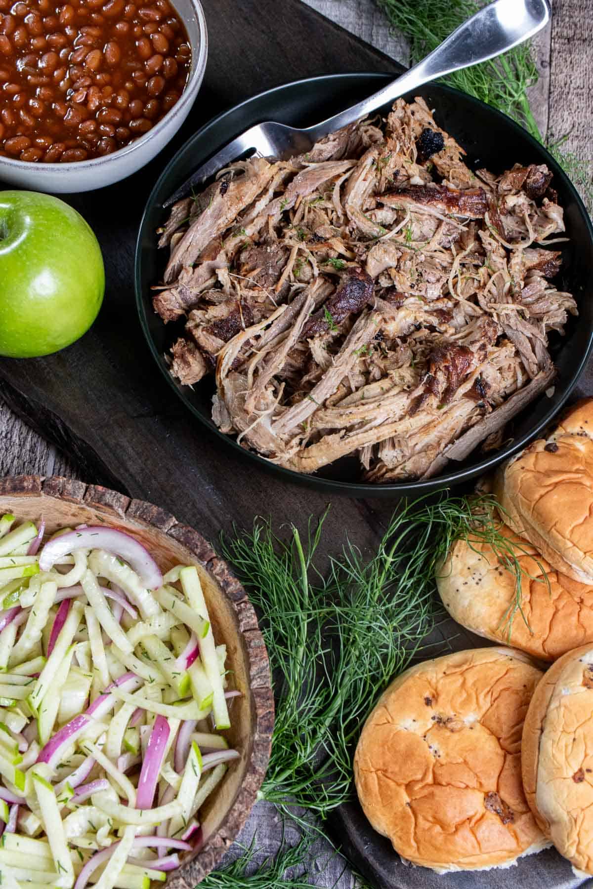 Angled view of cider braised pork shoulder, served with onion buns, fennel apple slaw, and baked beans.