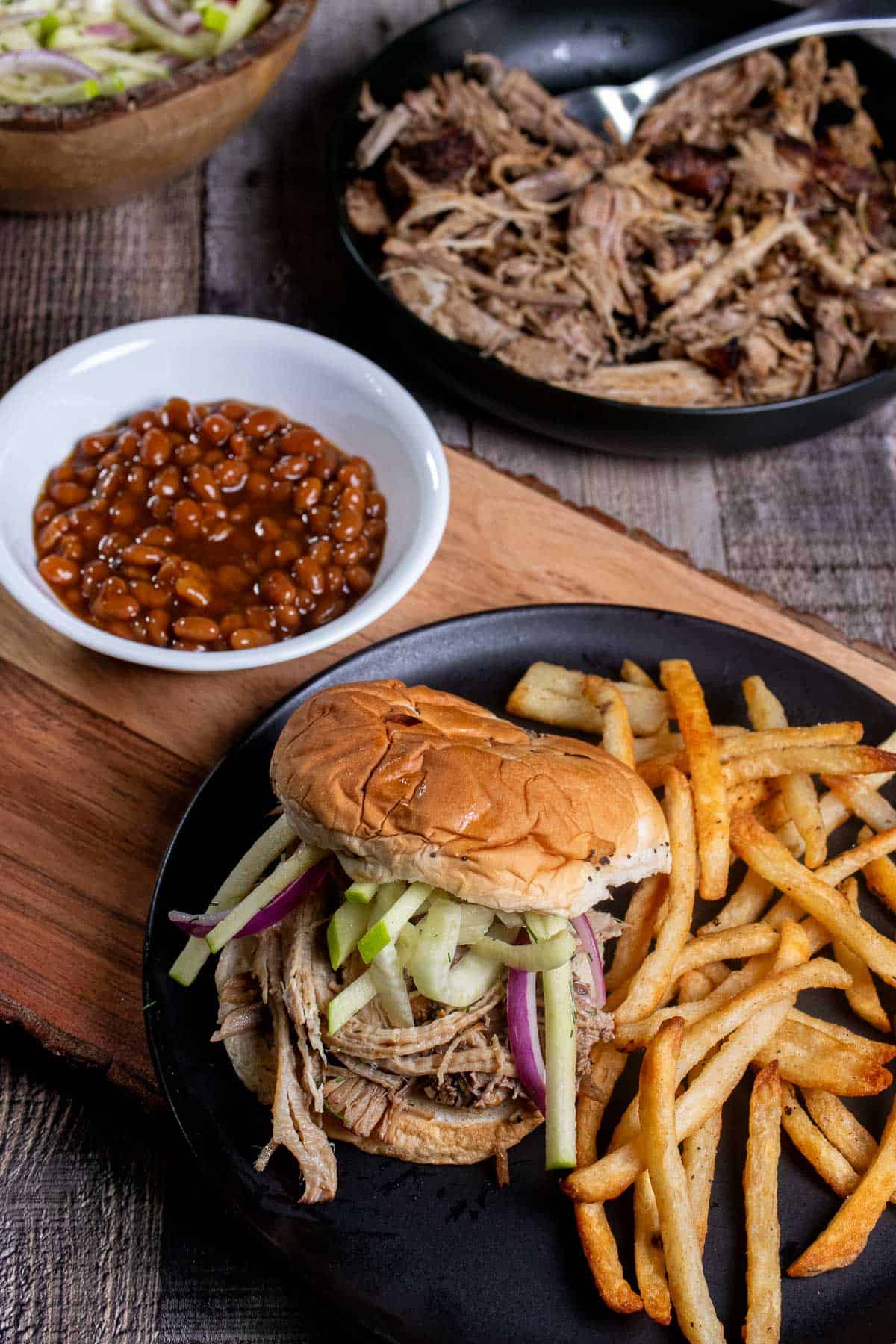 Cider braised pulled pork on an onion roll topped with fennel apple slaw and served with French fries on a black plate. 