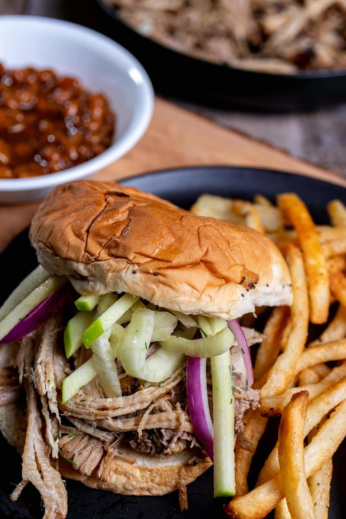 Cider braised pulled pork on an onion roll topped with fennel apple slaw and served with French fries on a black plate.