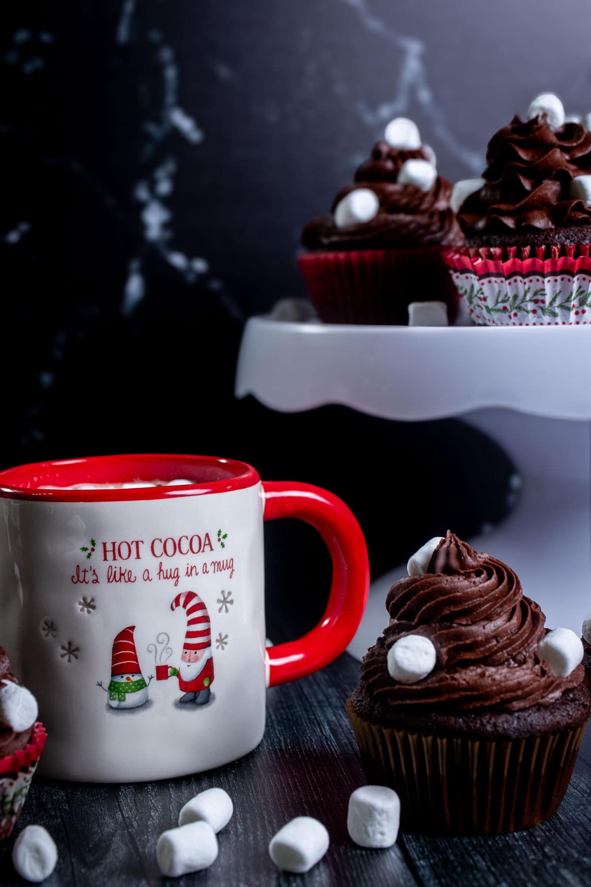 Wide shot of hot cocoa cupcakes next to a mug of hot chocolate with more cupcakes on a white cake stand behind them.