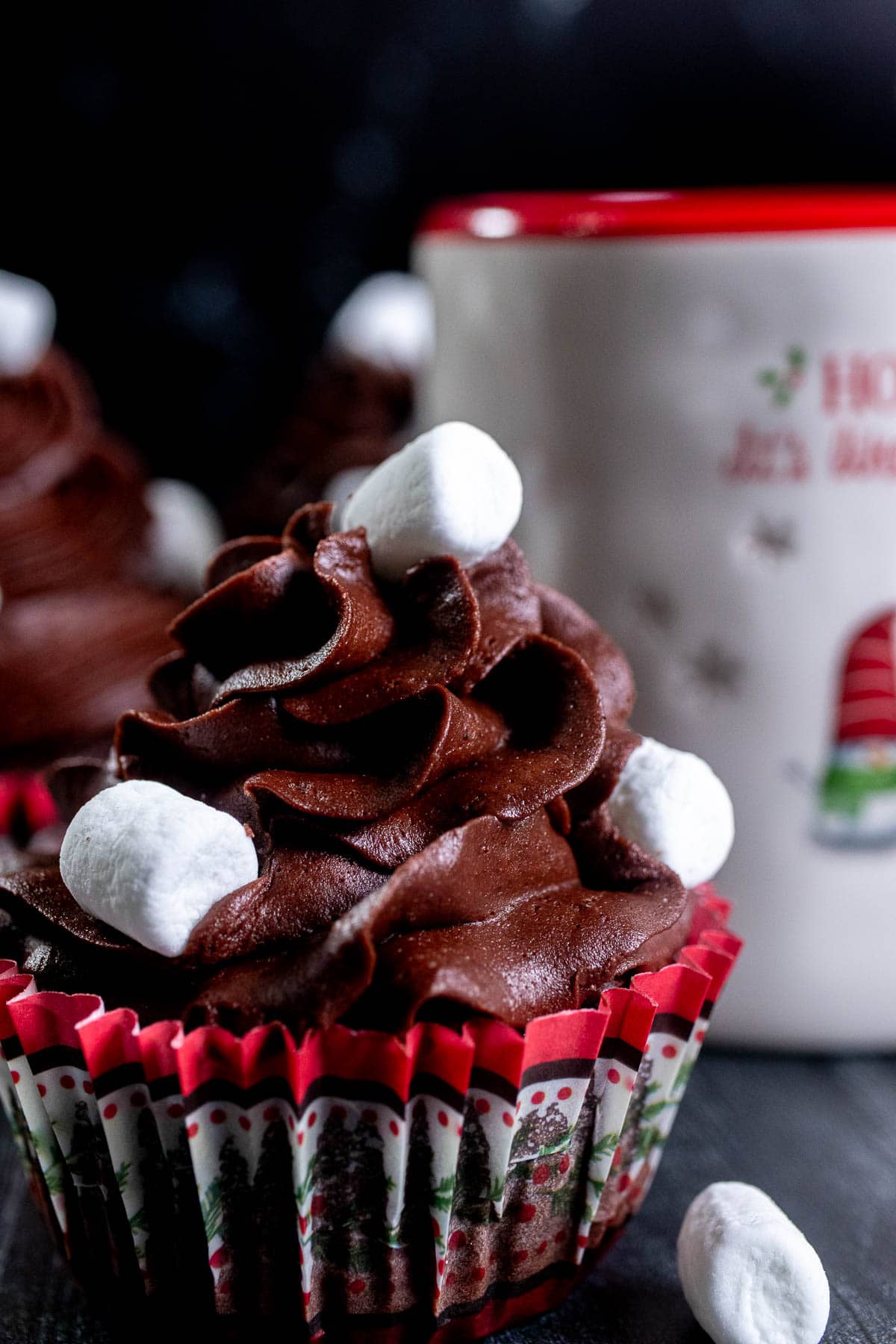 Close up of a hot chocolate cupcake with mini marshmallows on top and in front of a mug of hot chocolate.