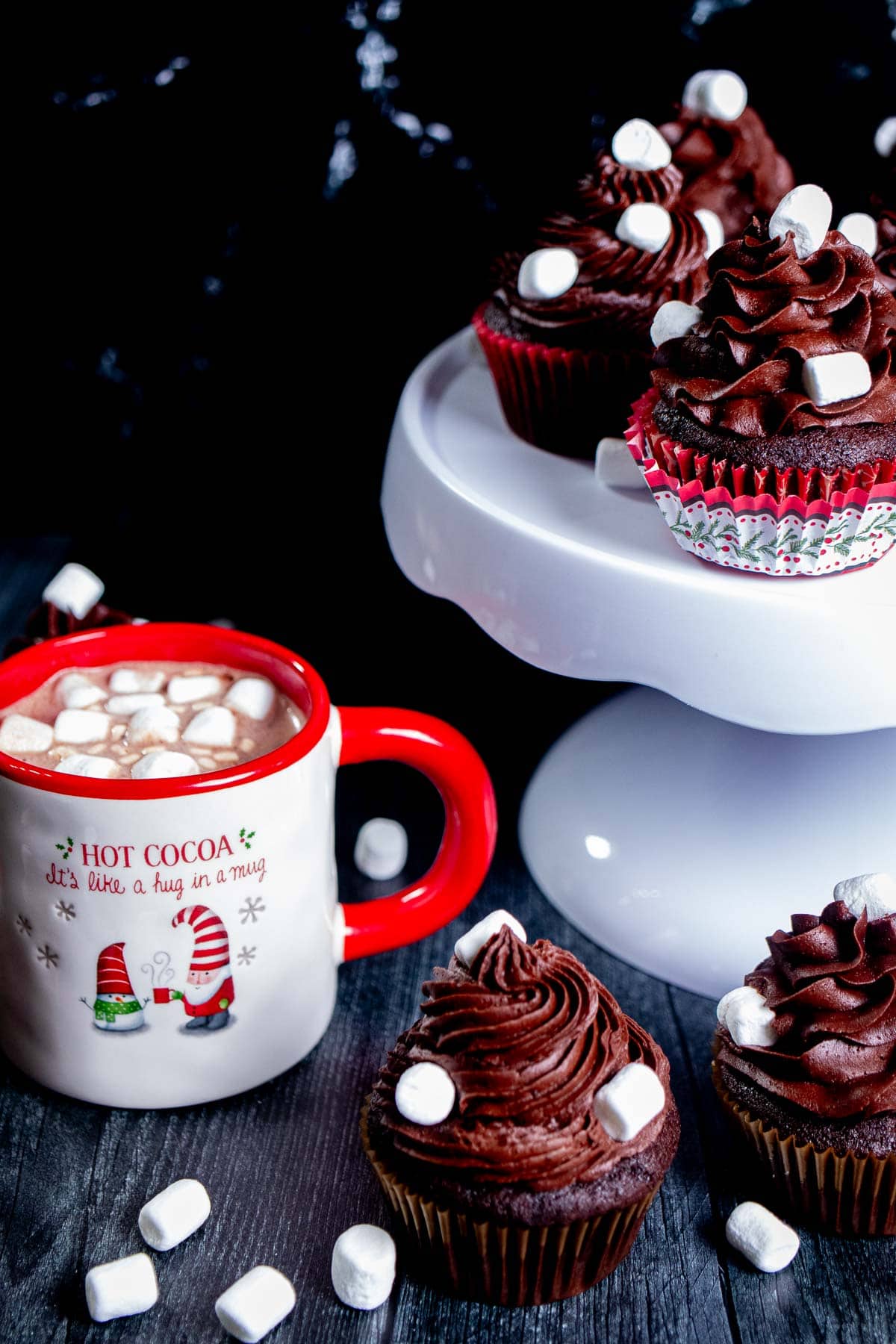 Angled view above a bunch of chocolate cupcakes on wood by a mug of hot chocolate with marshmallows and a white cake stand with more cupcakes on it.