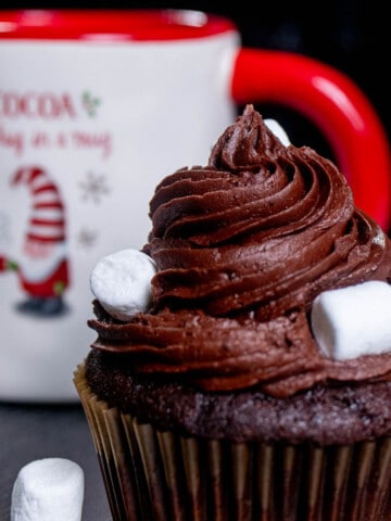 Close up of a hot cocoa cupcake with chocolate frosting in front of a Christmas cup of hot chocolate.