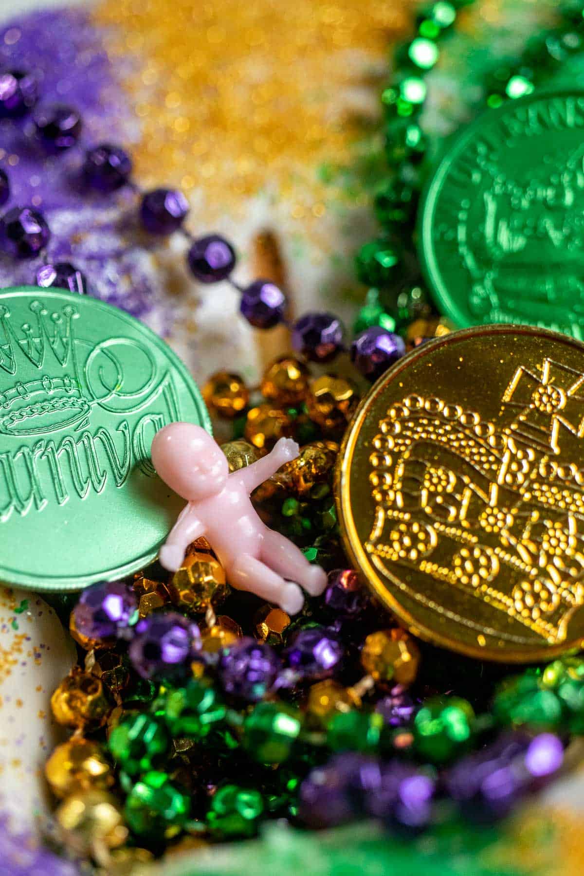 Close up view of the baby on top of a Louisiana king cake, nestled between Mardi Gras beads and coins.