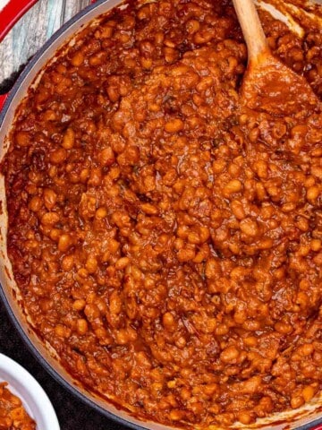 Large braising dish full of smoky BBQ baked beans with a wooden spoon in the corner.