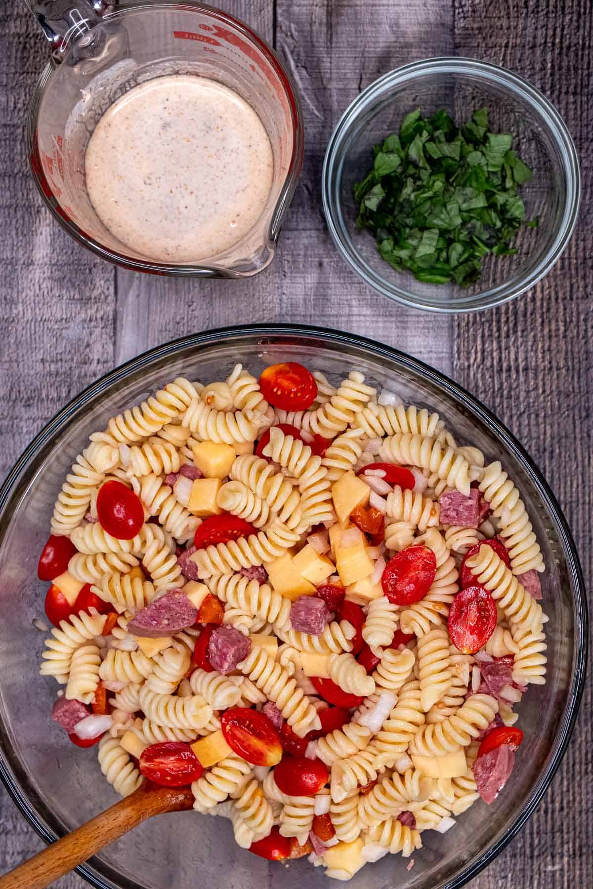 A large glass mixing bowl with pasta salad mixed together and above that a glass with the dressing and a bowl with fresh basil.