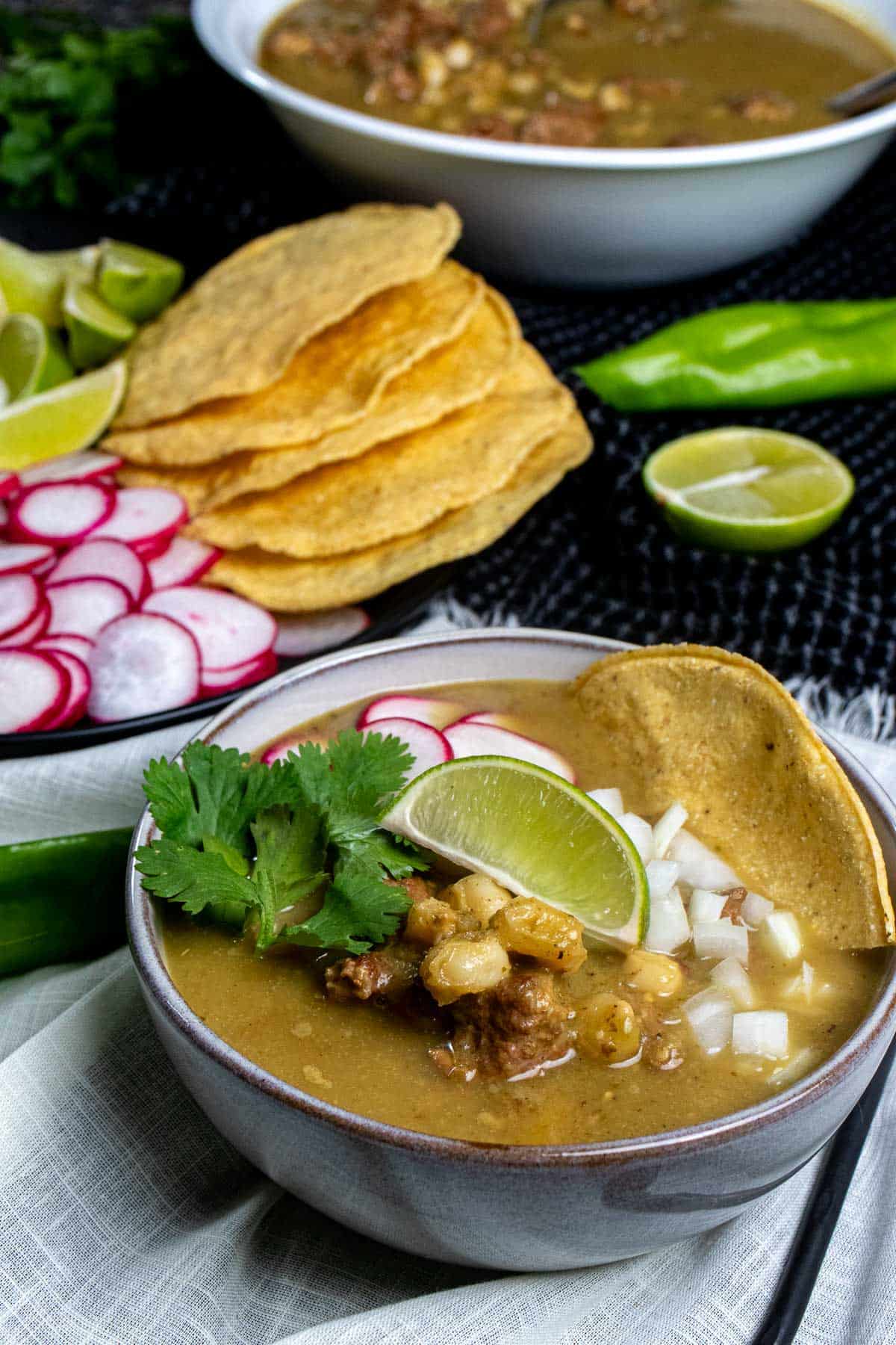 Pork pozole verde in a bowl topped with cilantro, onions, and radishes. More toppings and pozole served behind it.