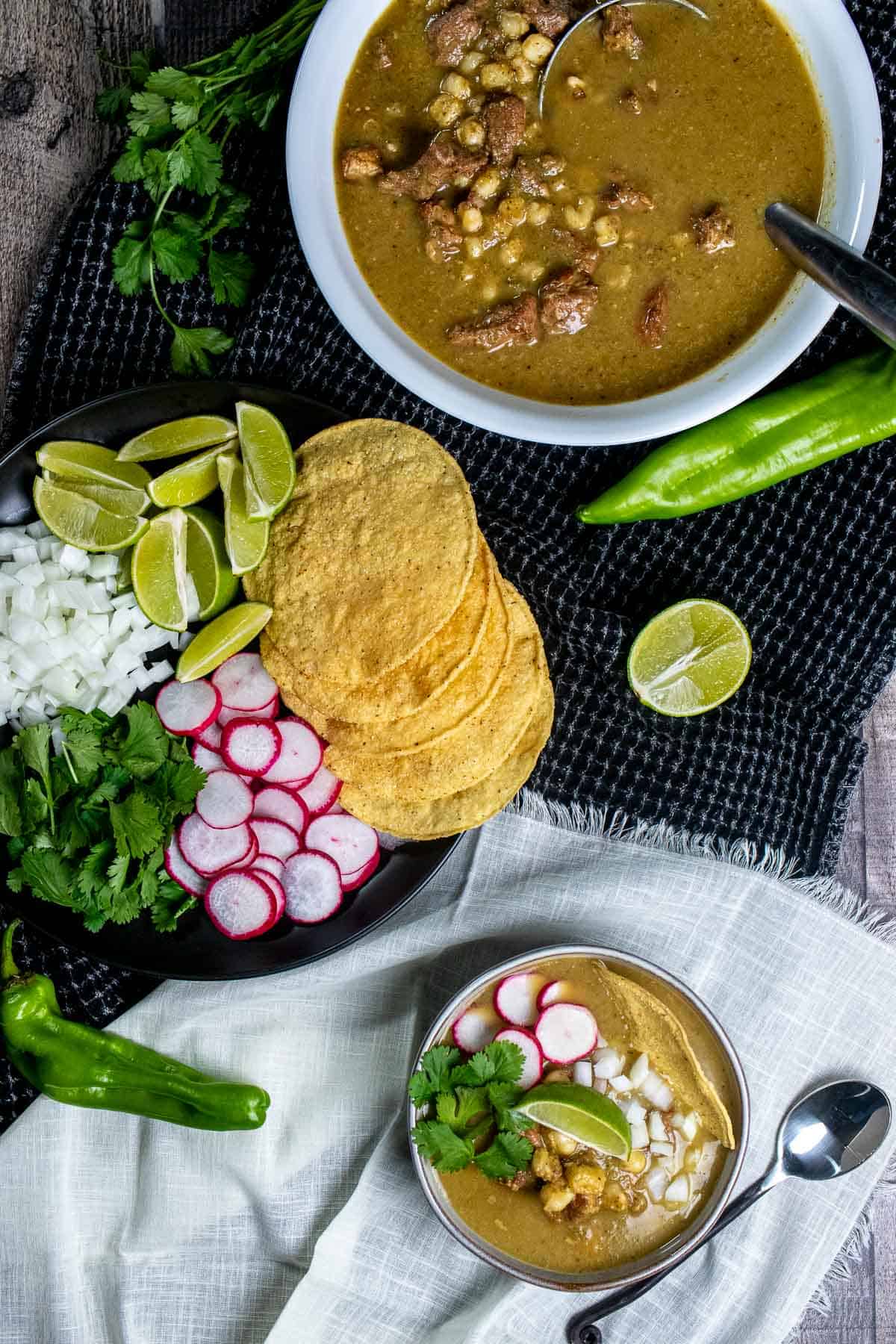 And overhead view of New Mexican pork pozole verde in a serving bowl, with a large plate of various toppings and a smaller bowl with pozole and toppings in it.