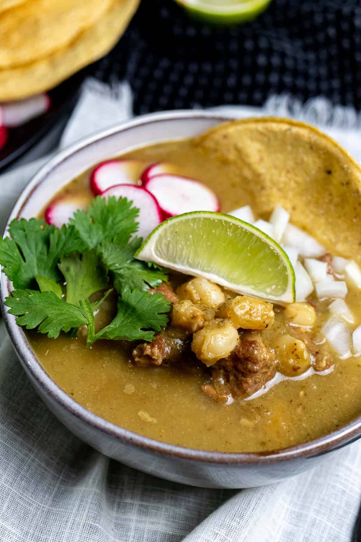 Pork pozole verde served in a bowl and topped with sliced radishes, diced onion, and cilantro with a tostado for dipping.