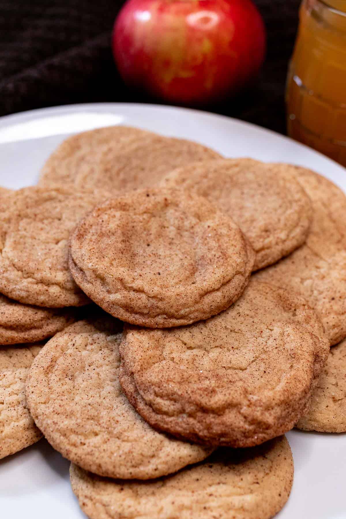 Apple cider snickerdoodle cookies piled onto a plate with an apple and a glass of cider behind them.