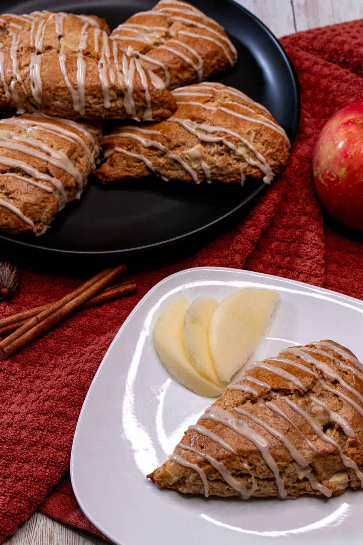 An apple cinnamon scone drizzled with maple cinnamon glaze with more on a plate behind it.