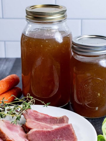 Two canning jars filled with homemade ham stock with a plate of carved ham in front of them.