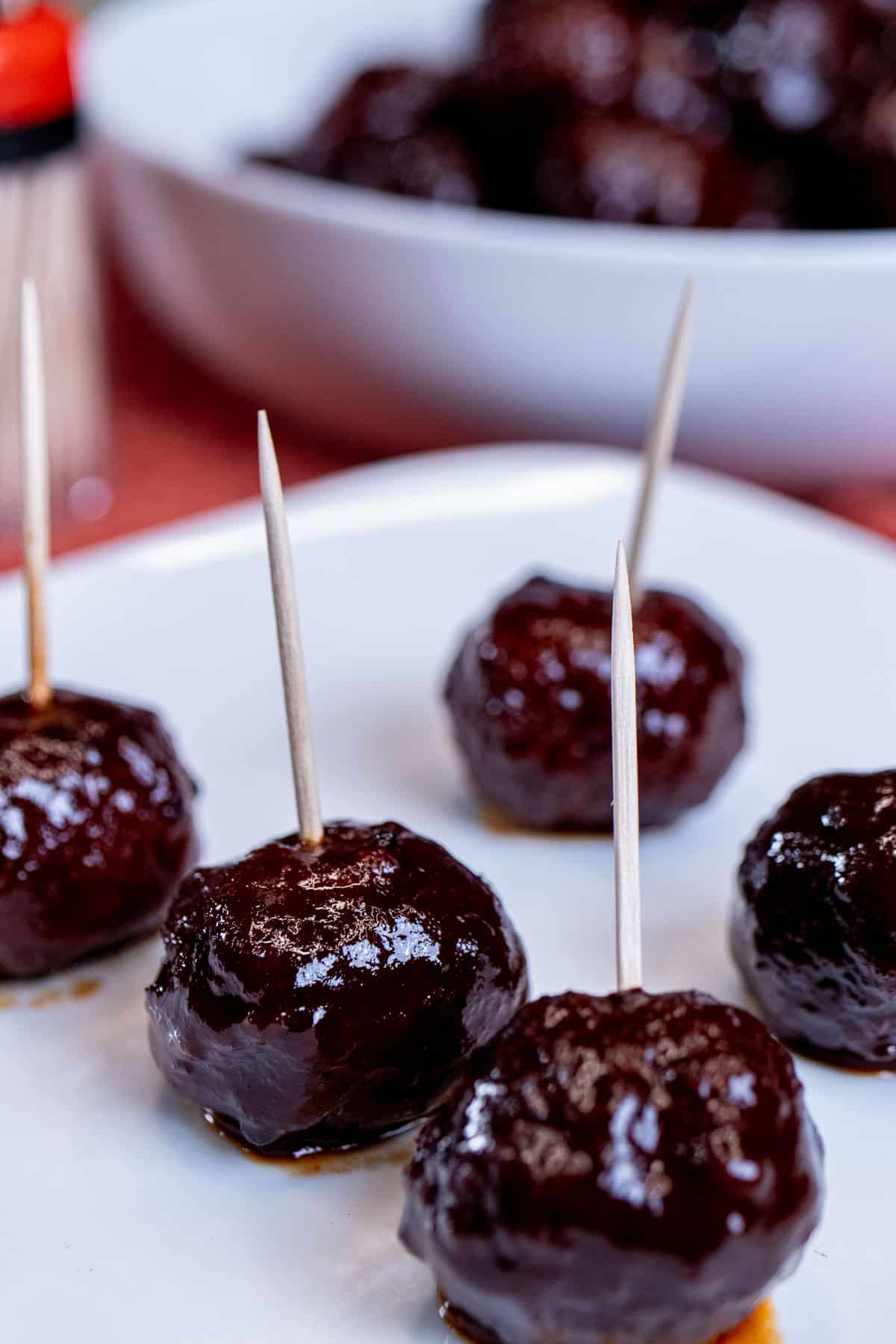 A plate of blackberry chipotle meatballs with toothpicks in them with more meatballs in the background.